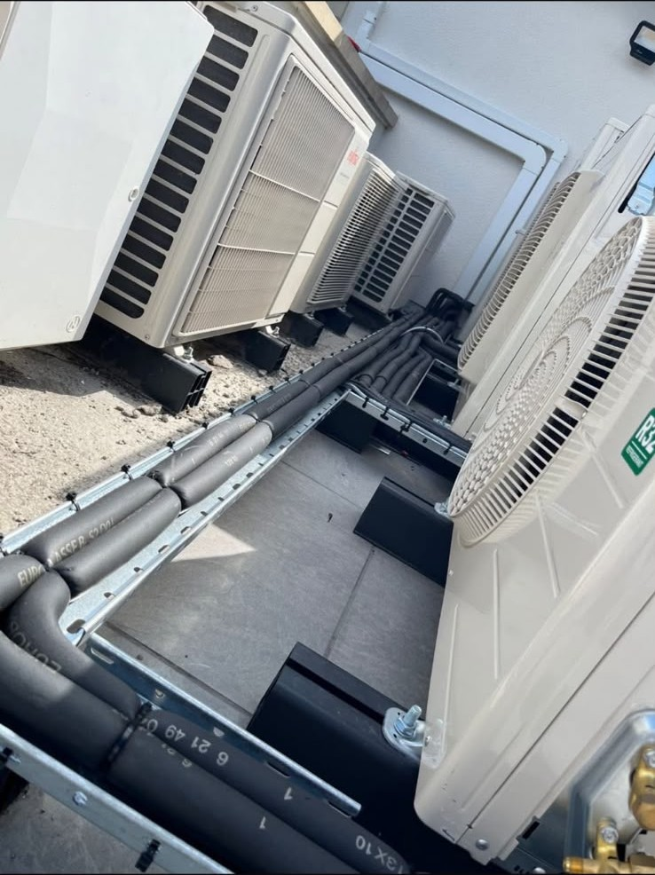 Row of outdoor condenser units on a commercial site with insulated refrigerant pipework on galvanised trays